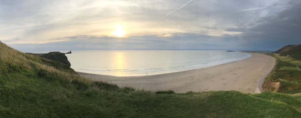 The sun is high in the sky and glistening off the sea. There is a panoramic view of a sandy beach surrounded by rolling hills.