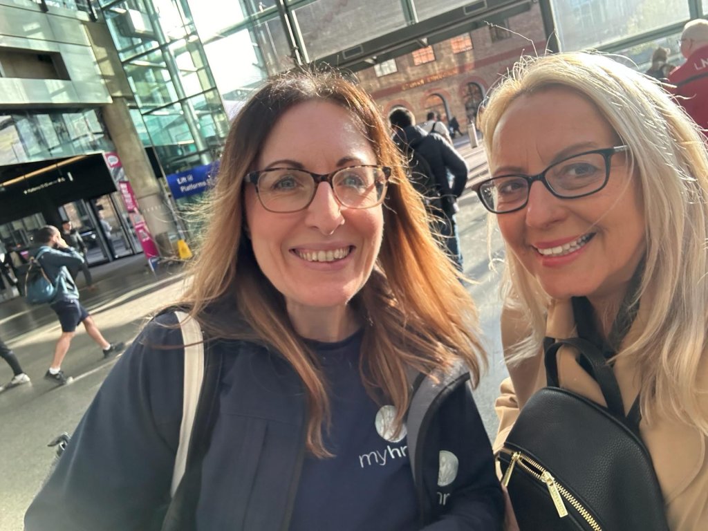 Rachael and Rebecca from My HR Hub are smiling as they stand in the center of a busy train station.