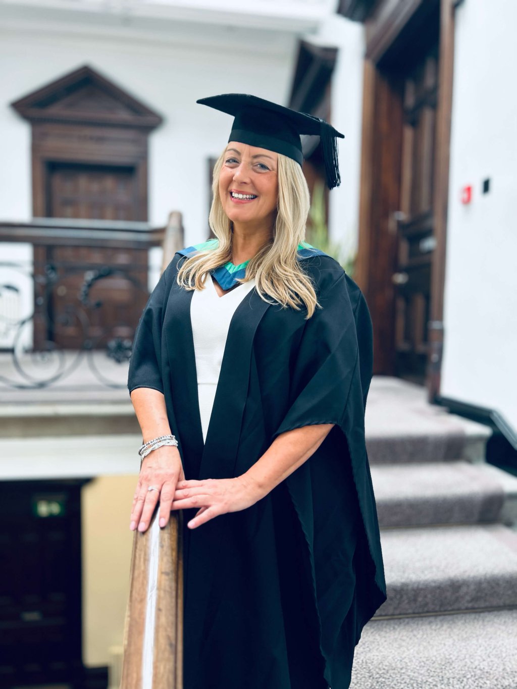 Rebecca Bull, founder of My HR Hub, is standing on a grand staircase in a graduation gown and hat.